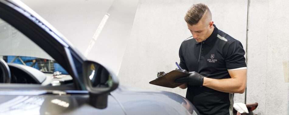 Mechanic in a Cox Auto uniform writing notes on a clipboard beside a car.