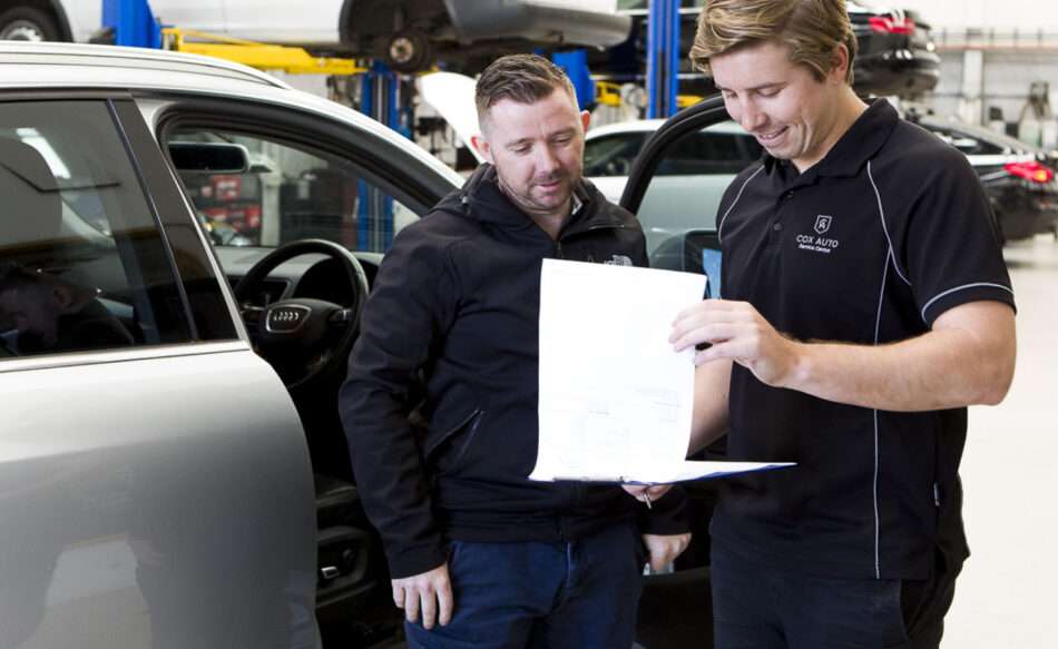 Two Cox Auto mechanics reviewing their service checklist, which includes battery checks.