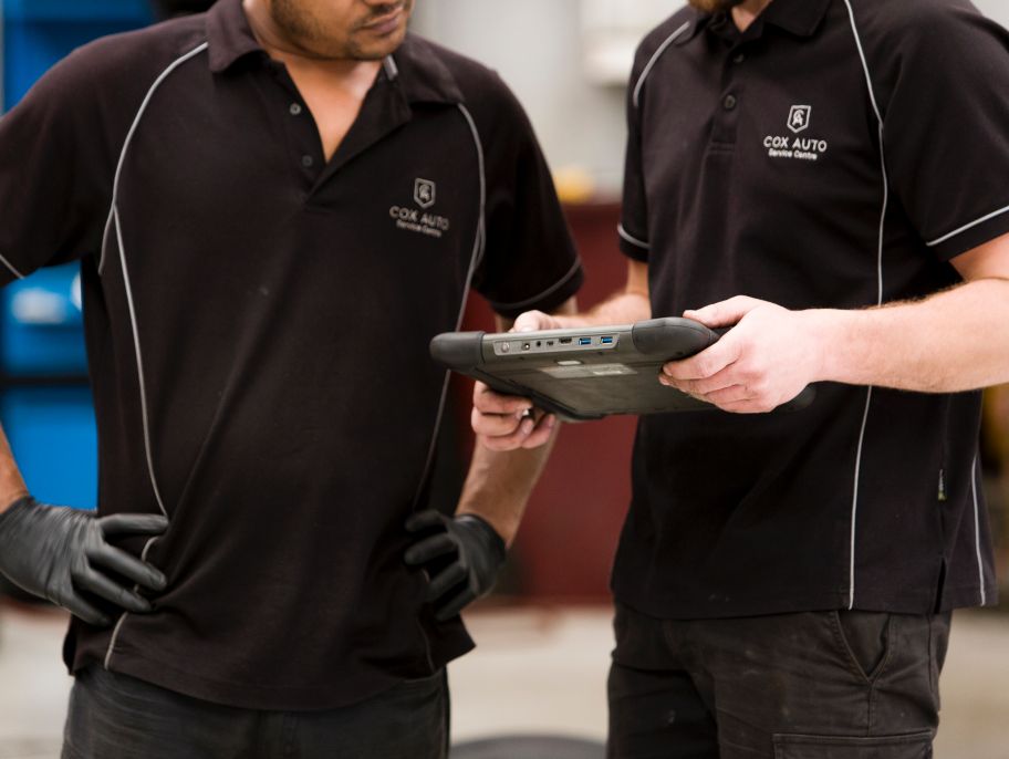 Two mechanics in Cox Auto uniforms examining data on a diagnostic tablet.
