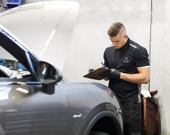 Mechanic in Cox Auto uniform inspecting a car and writing notes on a clipboard.