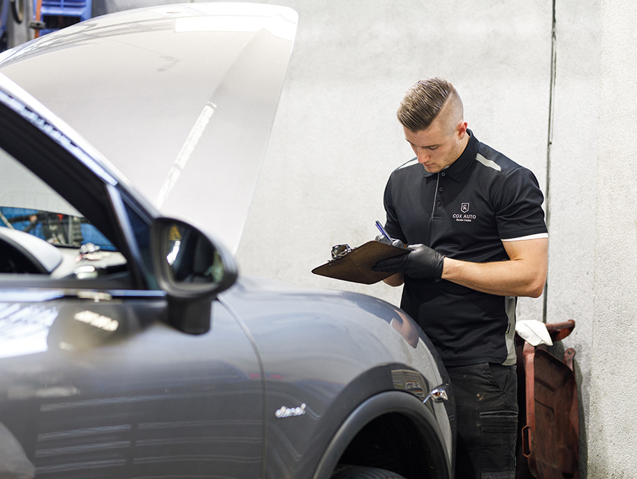 Mechanic in Cox Auto uniform inspecting a car and writing notes on a clipboard.