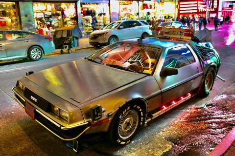 A replica DeLorean is parked on the side of a street at night, surrounded by city lights and urban surroundings.