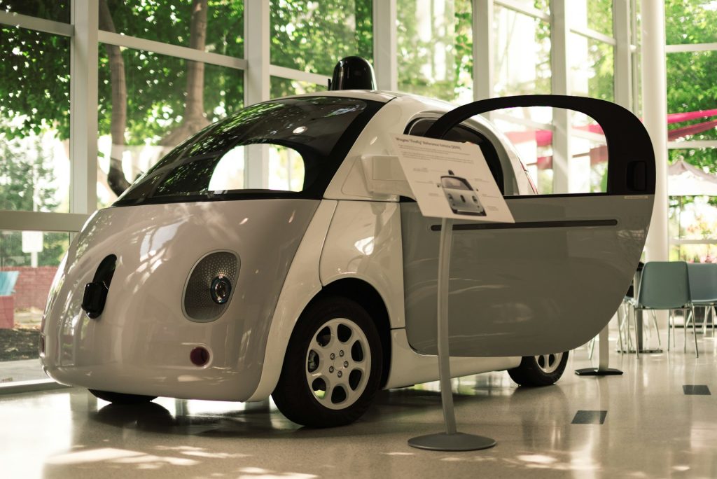 A small, white autonomous vehicle with an open front door is on display indoors, hinting at the future of driving. It's surrounded by large windows revealing trees outside, and a sign with information is positioned beside the vehicle.