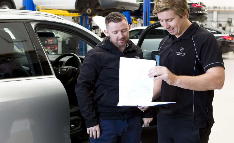 Service advisor at Cox Auto showing a vehicle report to a customer beside an Audi in the garage.