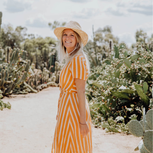 A young woman is walking through a cactus garden in the sunshine. She has blonde hair, wears a straw wide-brimmed hat and has turned back to the camera to smile. She wears a striped orange dress.