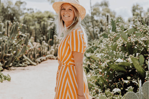 A young woman is walking through a cactus garden in the sunshine. She has blonde hair, wears a straw wide-brimmed hat and has turned back to the camera to smile. She wears a striped orange dress.