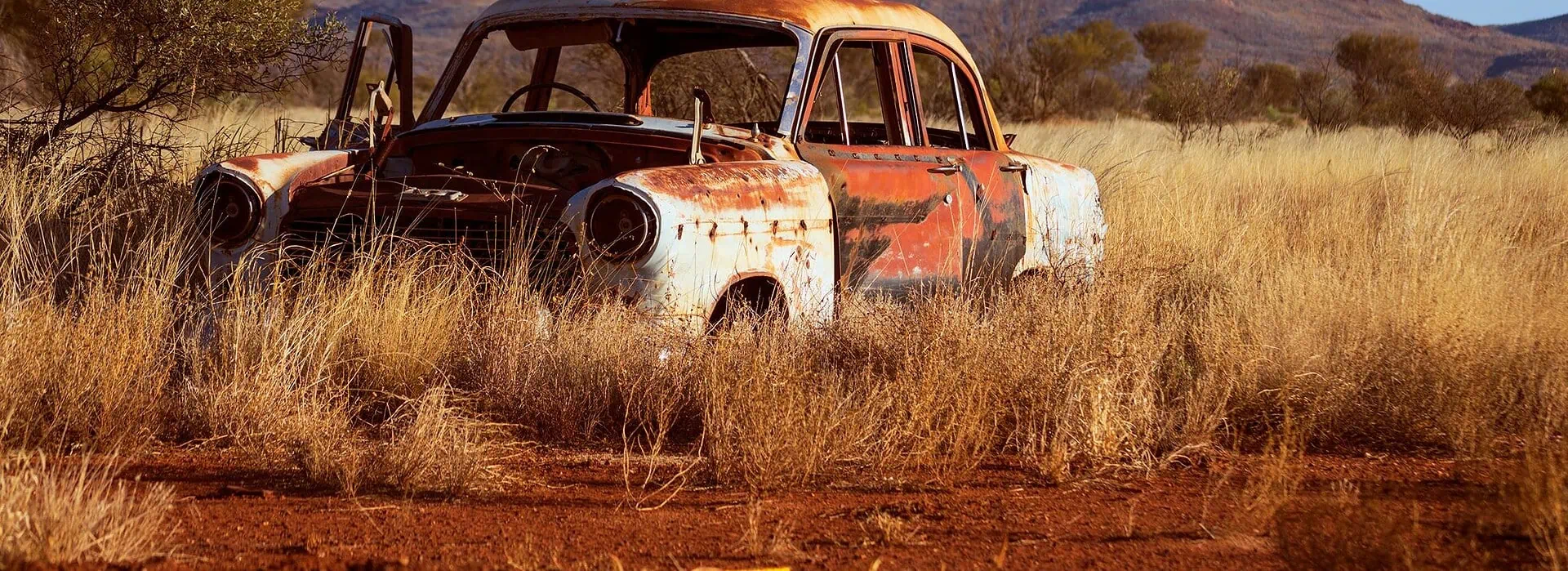 An old rusty car in the middle of a dry desert, surrounded by tall yellow grass with mountains in the background, depicting vintage vehicle decay. No car air-conditioning here!