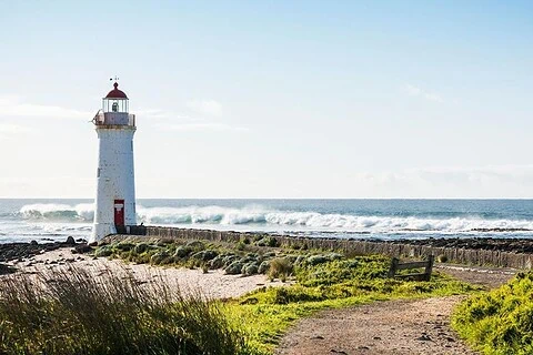 A lighthouse on a coastal landscape with a walking path and greenery, overlooking the ocean under a clear sky.
